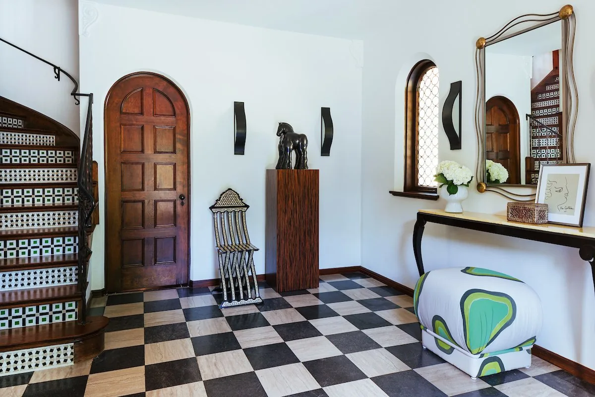 Happy Hollow foyer, black and white checkerboard marble floor, arched mahogany entry door, tiled stair risers, and sculptural horse on a pedestal. Studio Eckström