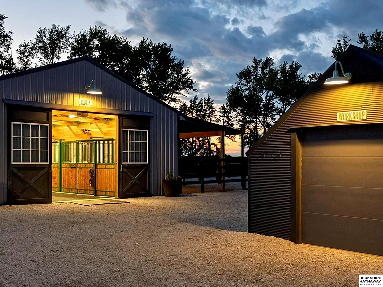 Equestrian stable interior entrance, cedar wall paneling, custom green powder-coated steel stall gates, and brick aisle at dusk. Country Equestrian, Studio Eckström