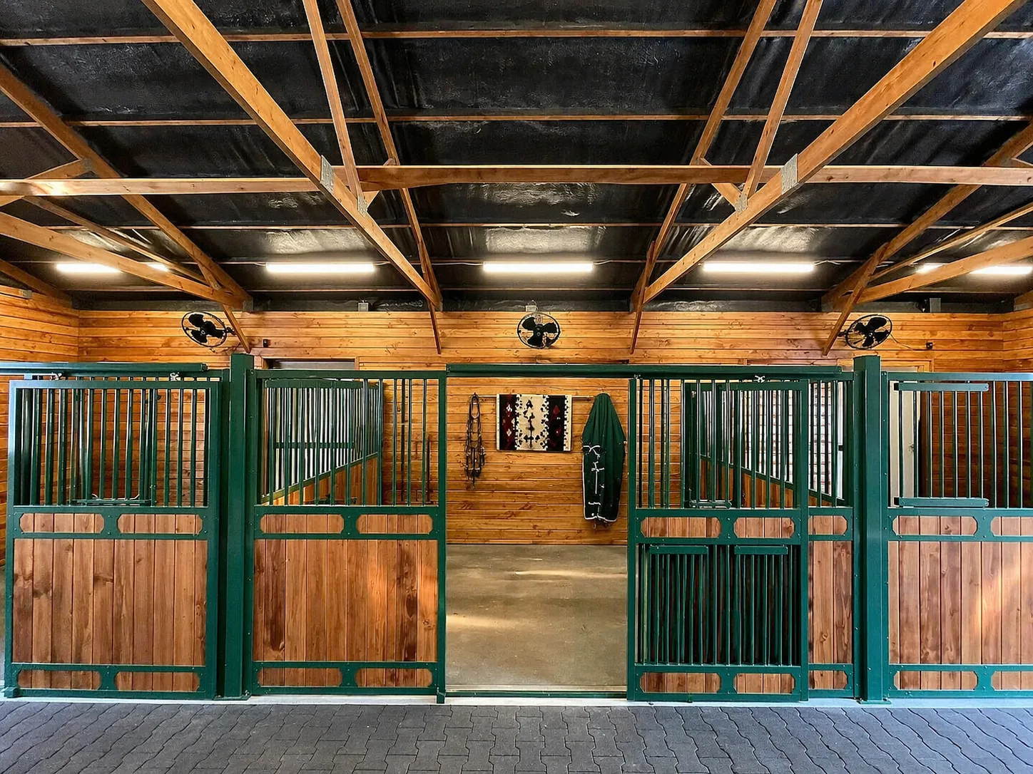Equestrian stable stalls, custom green steel stall gates, cedar paneling, exposed timber roof, and brick aisle. Country Equestrian, Studio Eckström