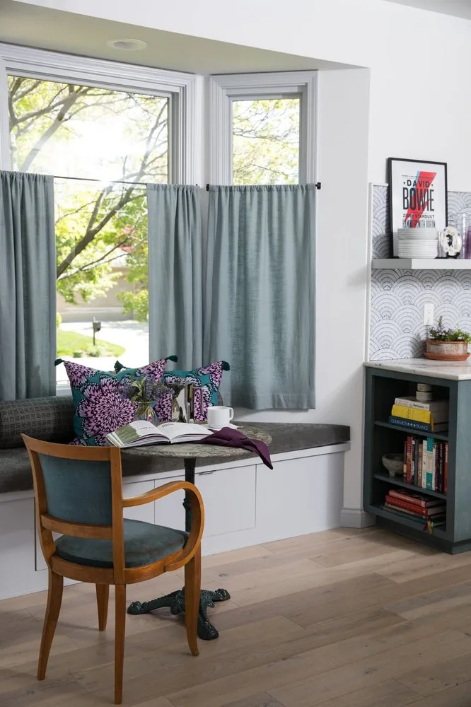 The Regency Refresh kitchen window seat, grey upholstered storage bench with floral pillows, bistro table with teal velvet chair, and grey linen curtains. Studio Eckström Omaha