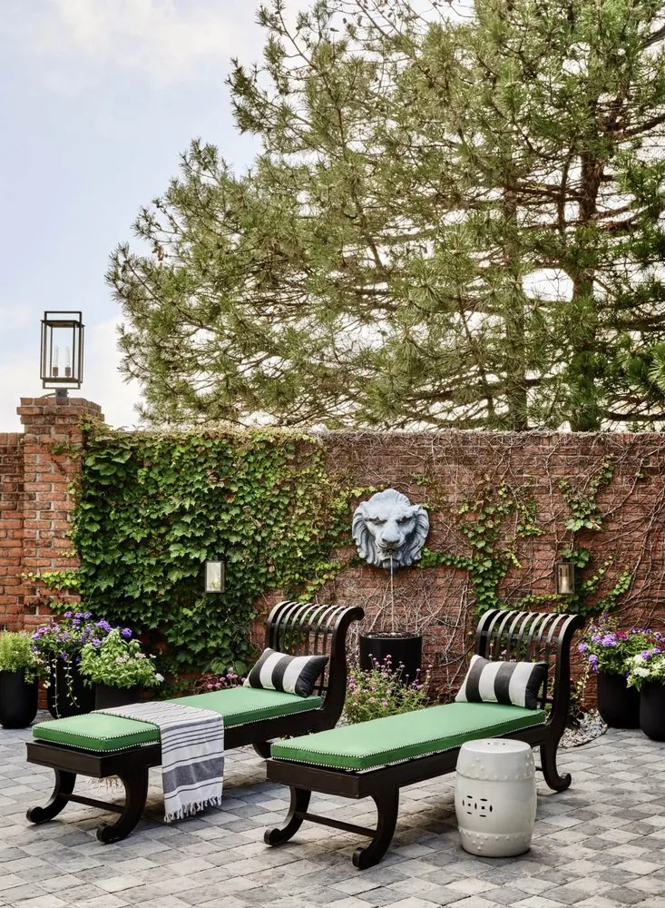 The Architectural Digest Townhome private courtyard, black iron Regency chaise longues, ivy-covered brick wall with stone lion fountain, and garden lanterns. Studio Eckström 2023