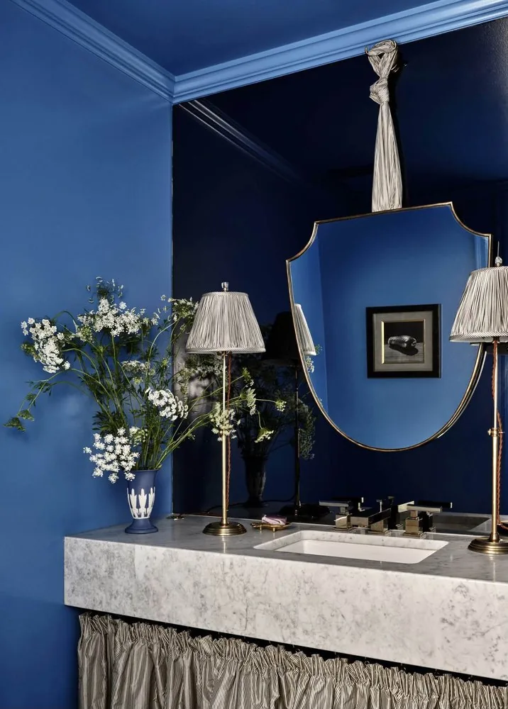 The Architectural Digest Townhome powder room, deep cobalt lacquered walls and ceiling, shield mirror, and marble vanity with ticking stripe skirt. Studio Eckström 2023